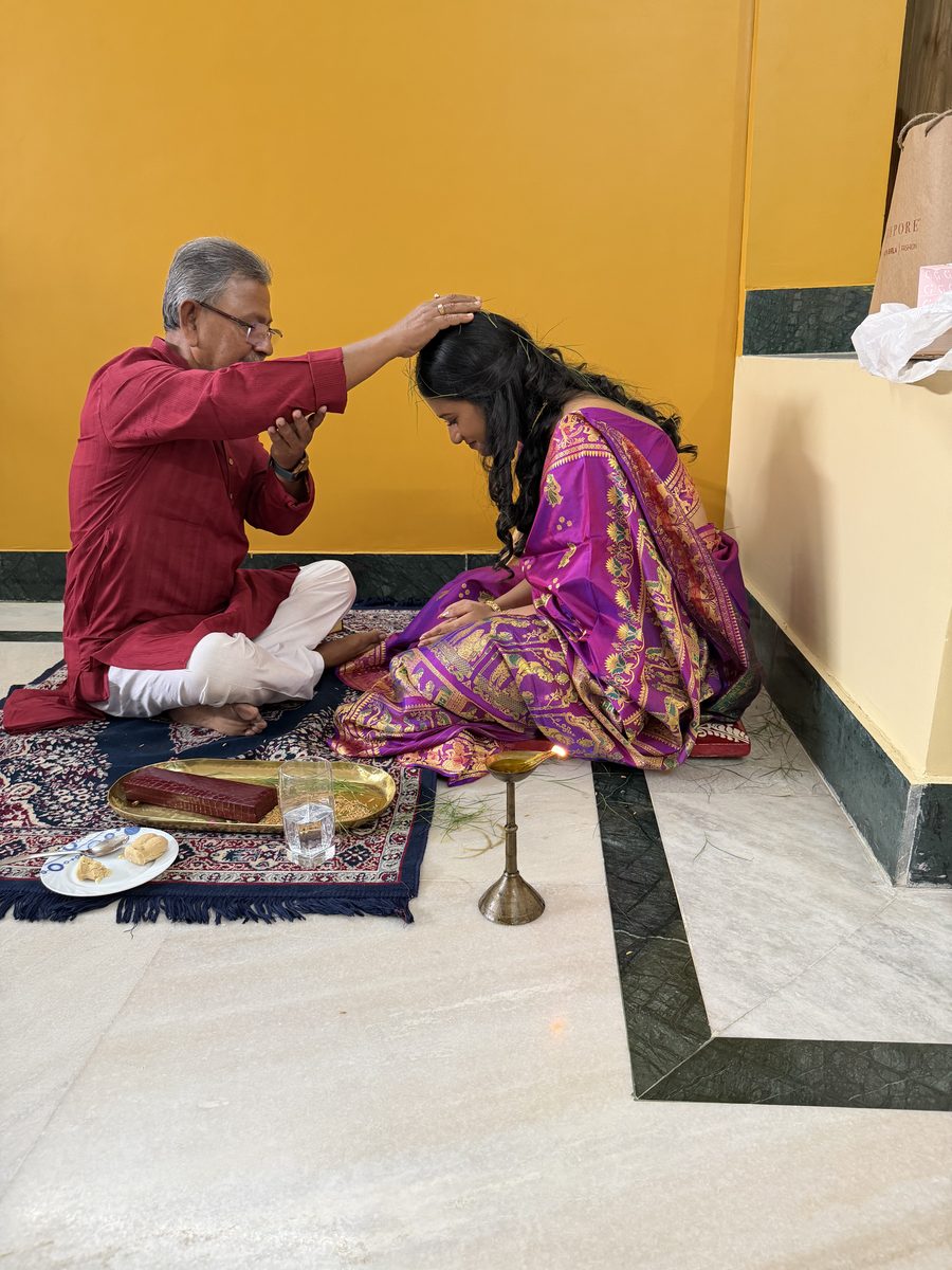 Elderly man in maroon kurta blessing Rituparna seated on the mat