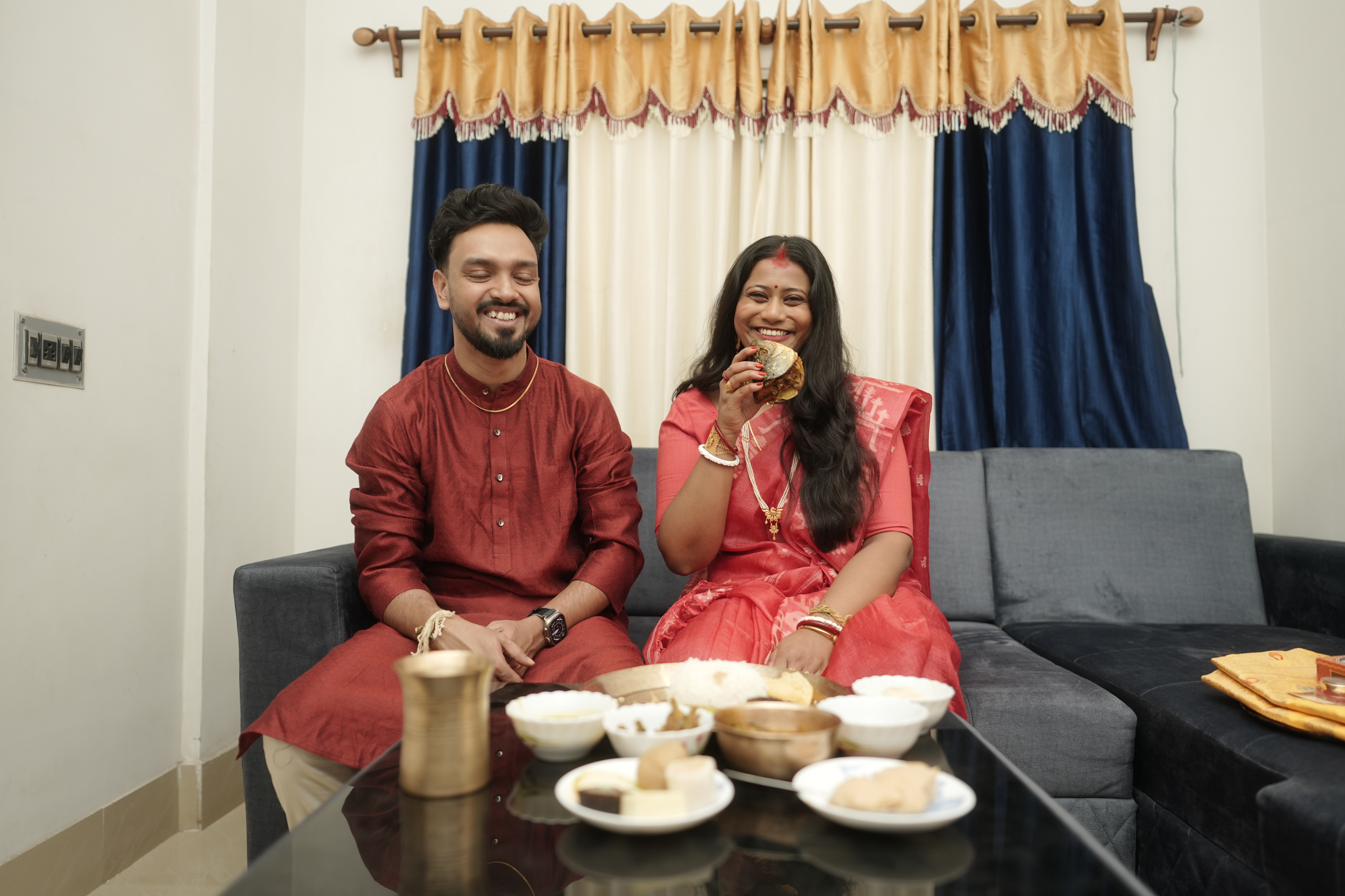 Rituparna and Shoumo sitting together on the couch, laughing while eating from the thali
