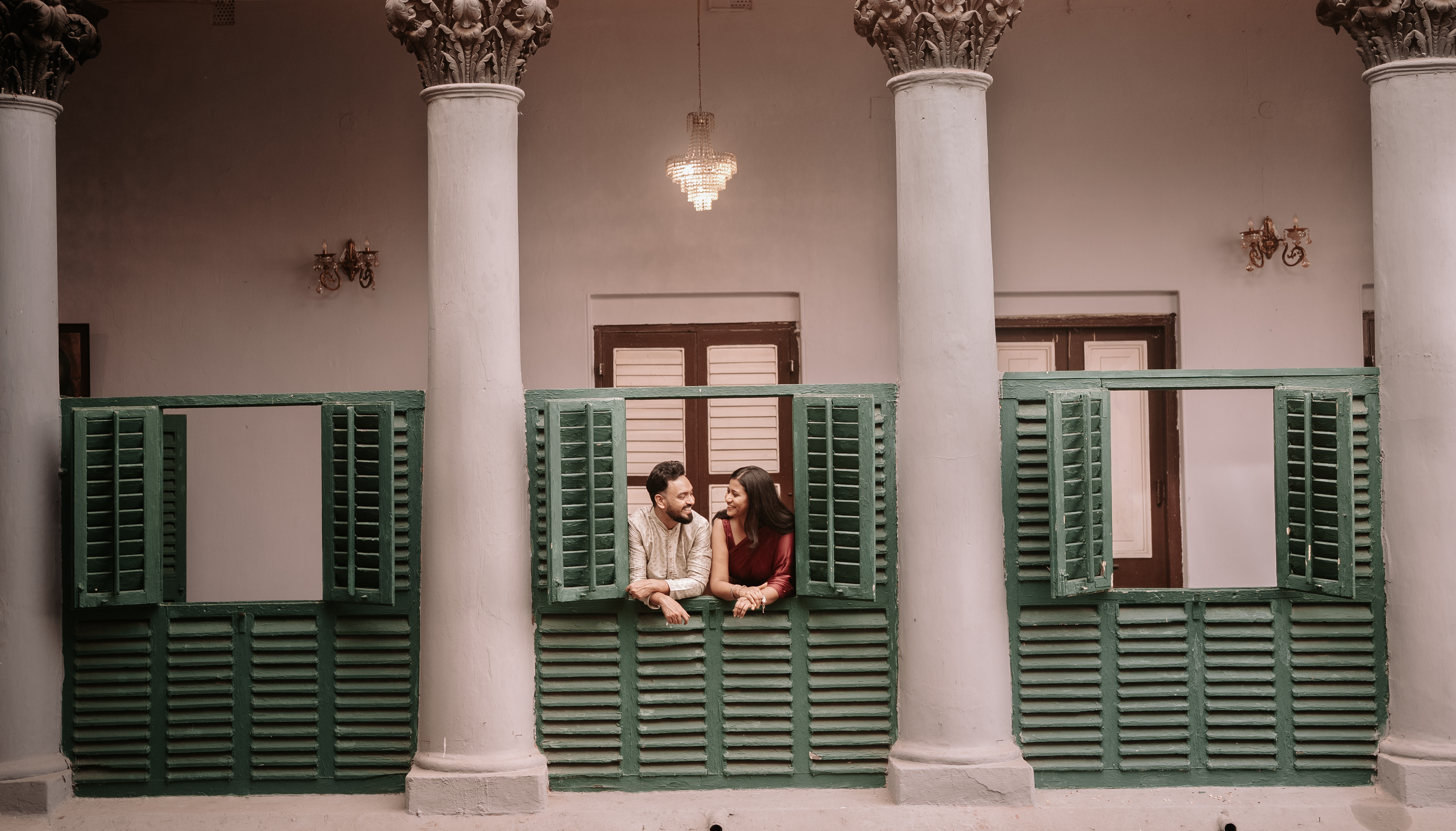 Wide shot of Rituparna and Shoumo playfully posing before the grand haveli facade