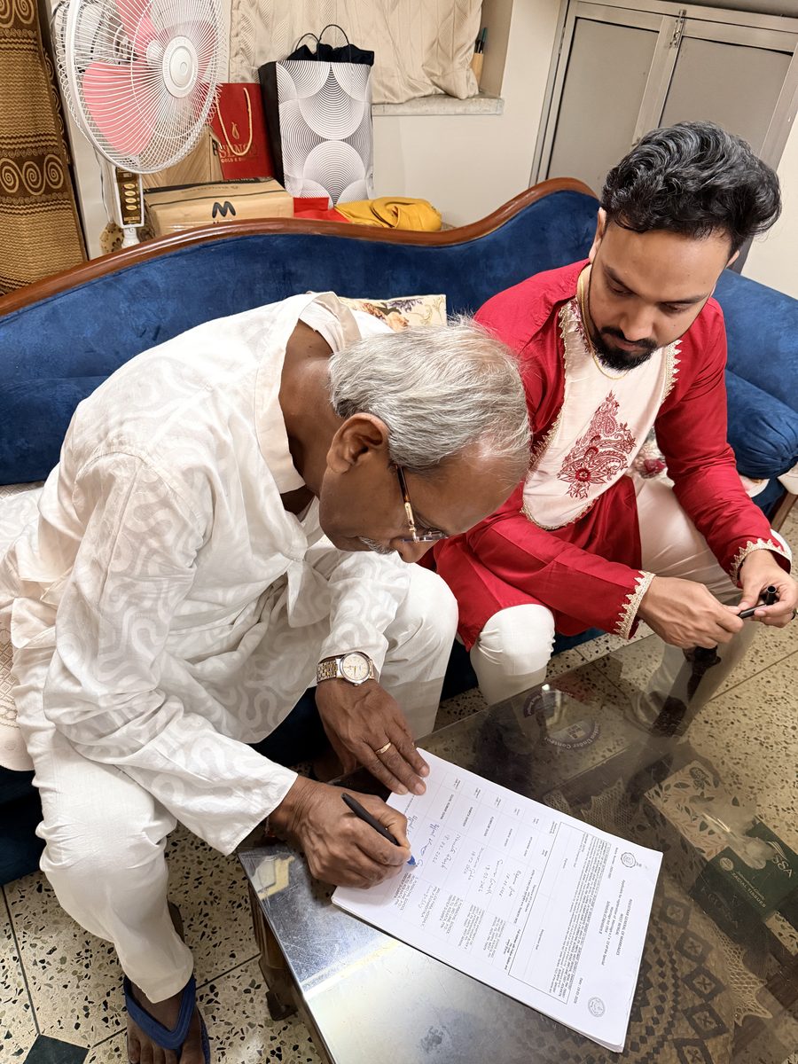 Elder man bending over to sign papers alongside Shoumo in red kurta